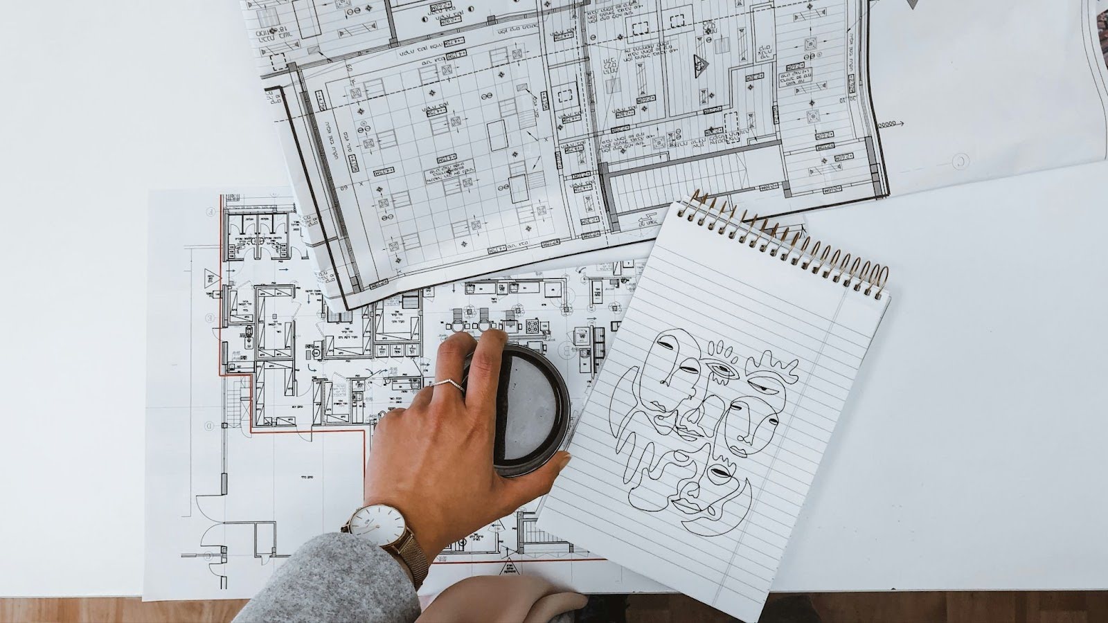 photo of several sheets of building plans and a sketchbook on a desktop with a hand resting on a cup of coffee in the center