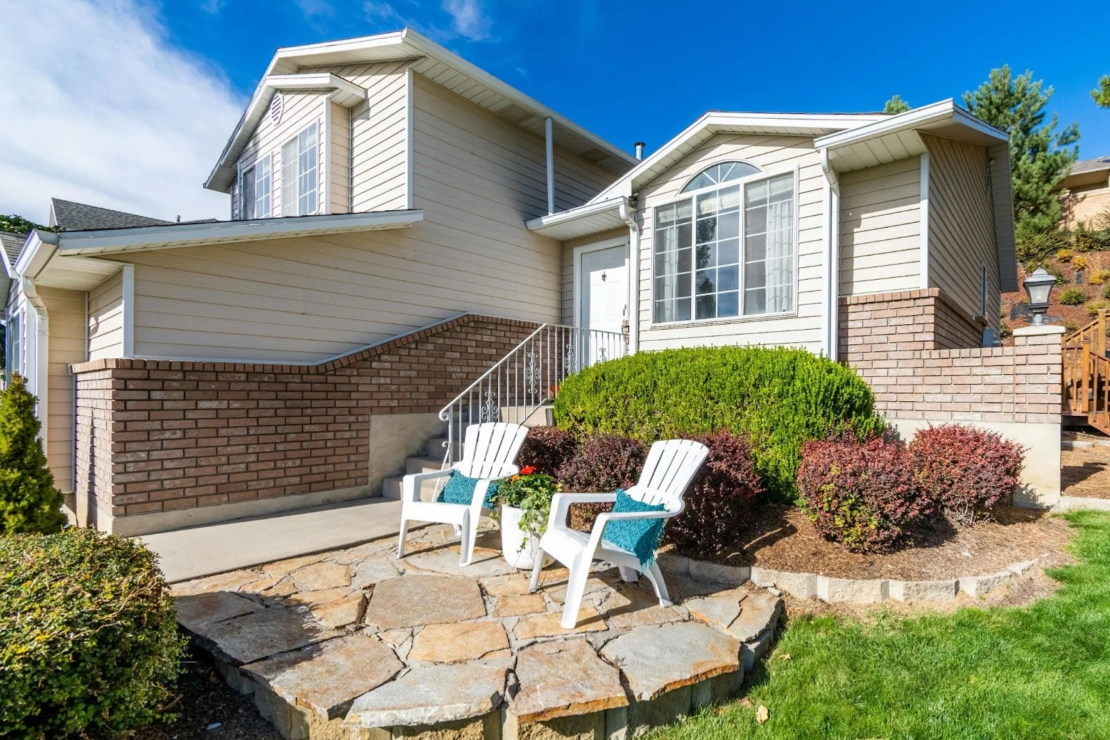 picture of a beige split level home featuring a stone paved patio with two white adirondack chairs and neatly manicured landscaping