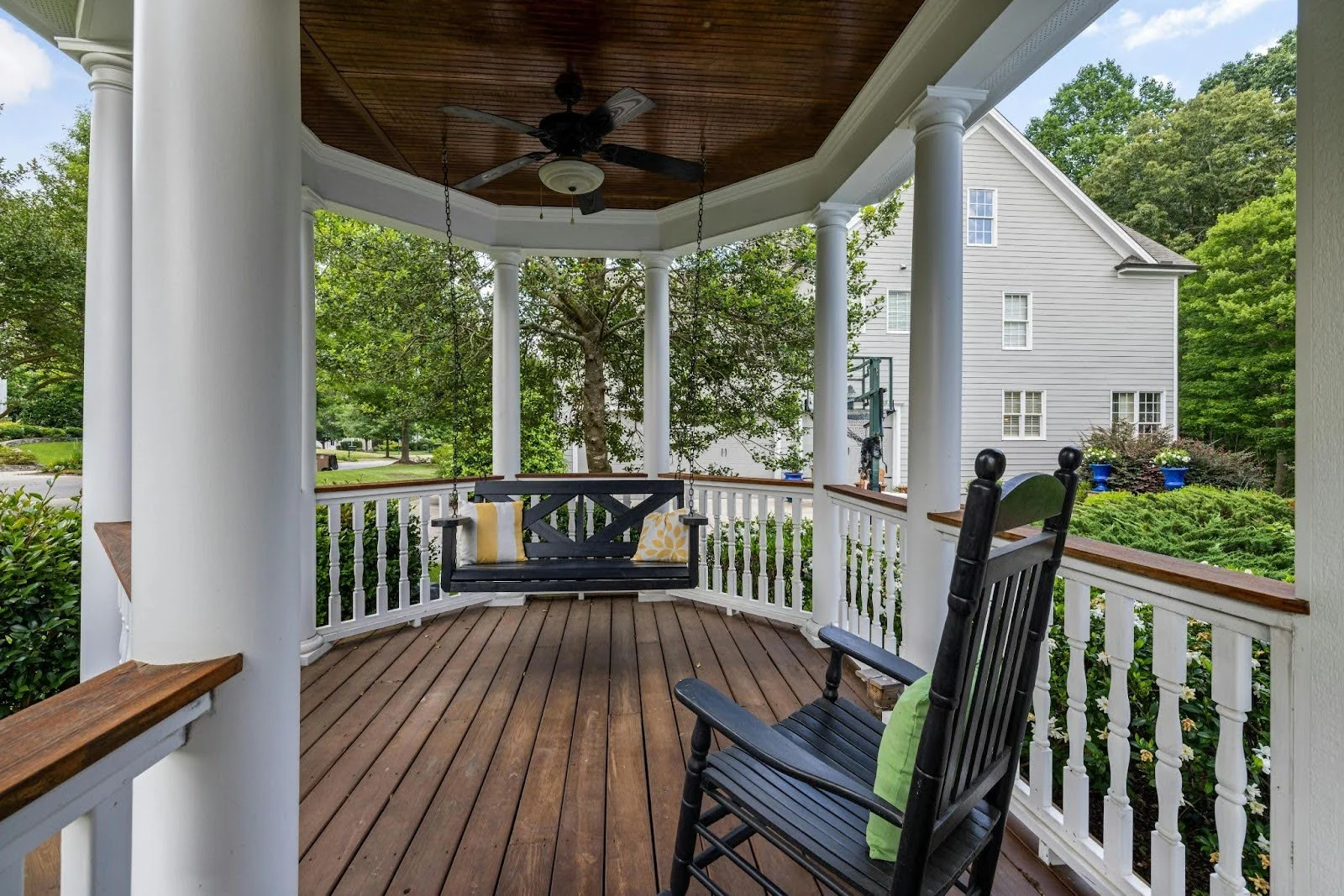 picture of a spacious white porch with circular columns features a dark wood ceiling a hanging porch swing and a black rocking chair overlooking a green lawn