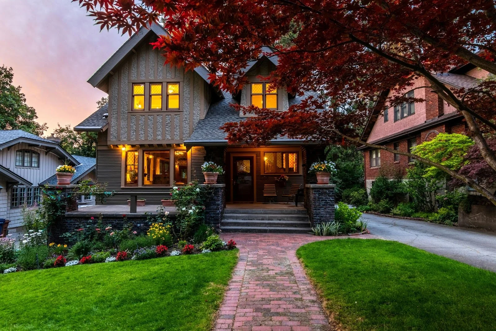 picture of a two story craftsman style home with lit windows and a brick paved walkway framed by a vibrant red maple tree at dusk