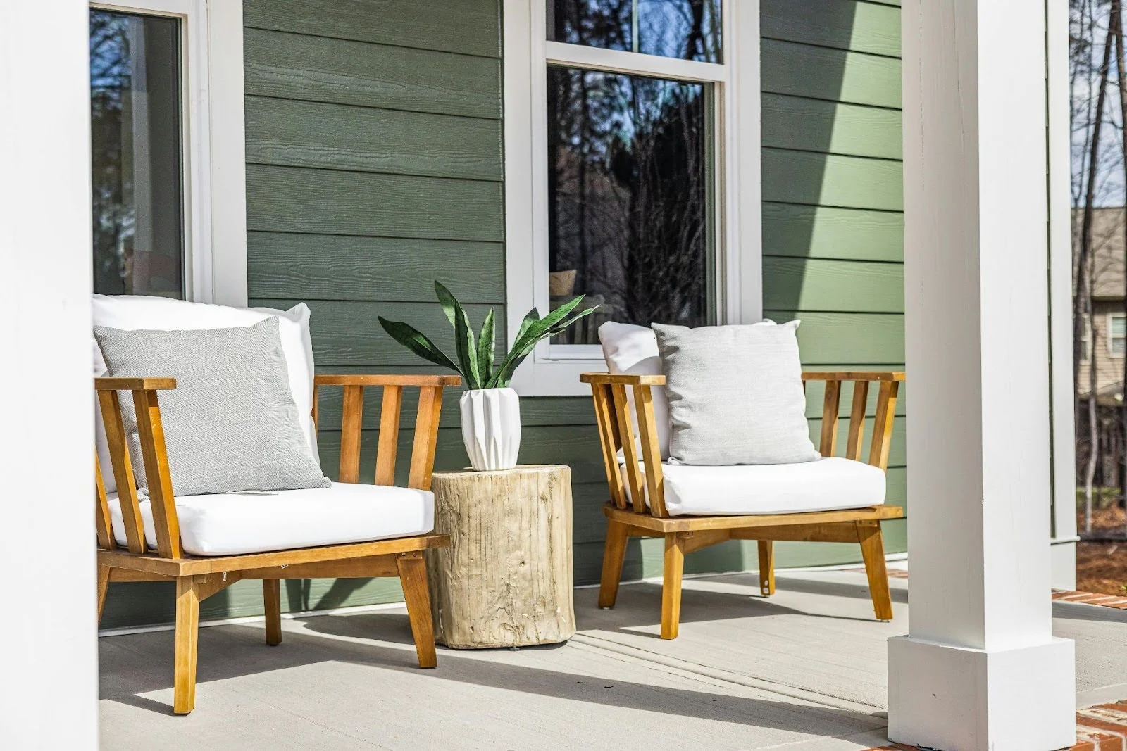 picture of two wooden armchairs with white cushions and gray pillows sitting on a concrete porch next to a natural wood stump side table with a plant on it against green siding