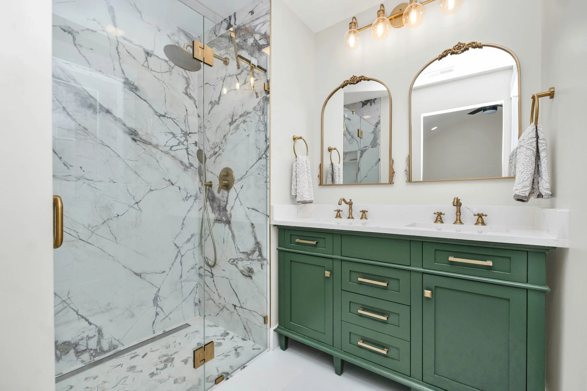 photo of a bathroom remodel with a green vanity and gold fixtures with white granite below mirrors and gold light fixtures and next to a white marble stand up shower