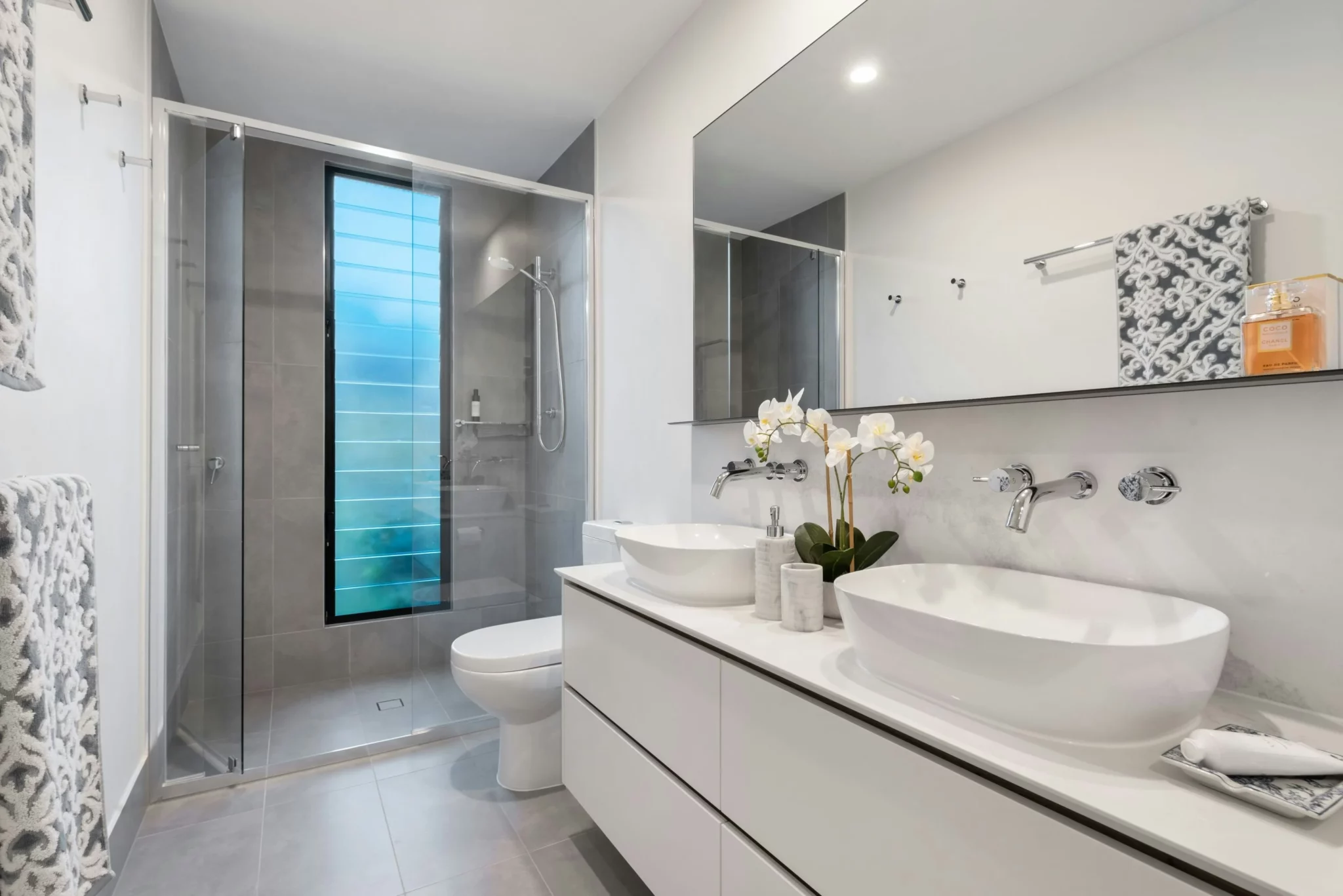photo of a modern bathroom with a large mirror and a white vanity with two bowl sinks next to a toilet and stand up shower