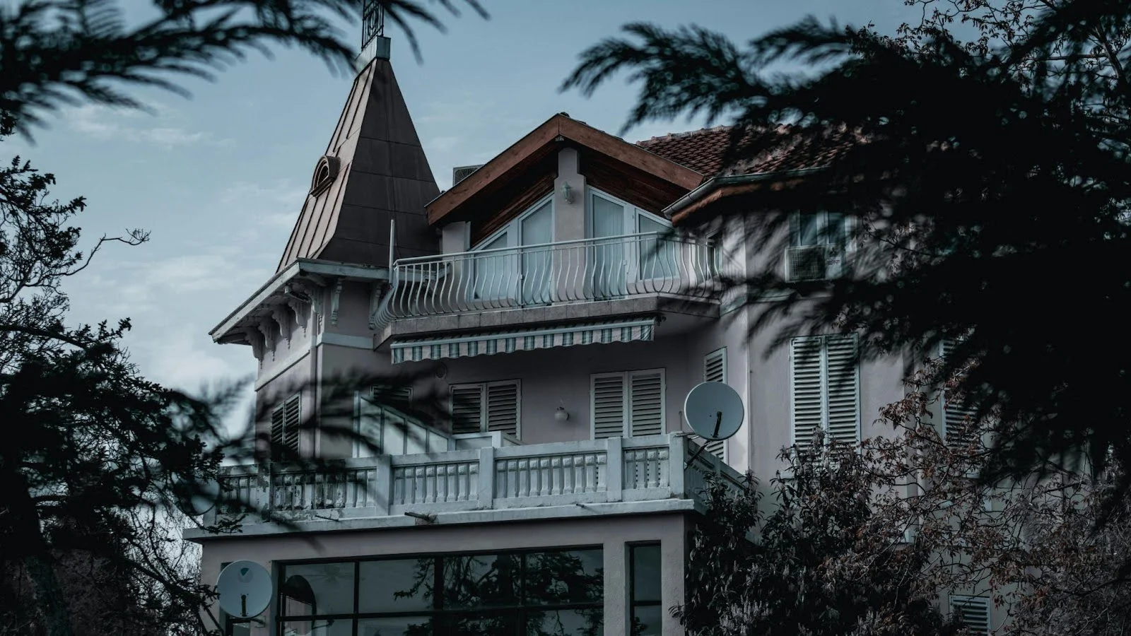 picture of a large pink european style estate with a pointed tower roof and multiple white balconies framed by dark tree branches