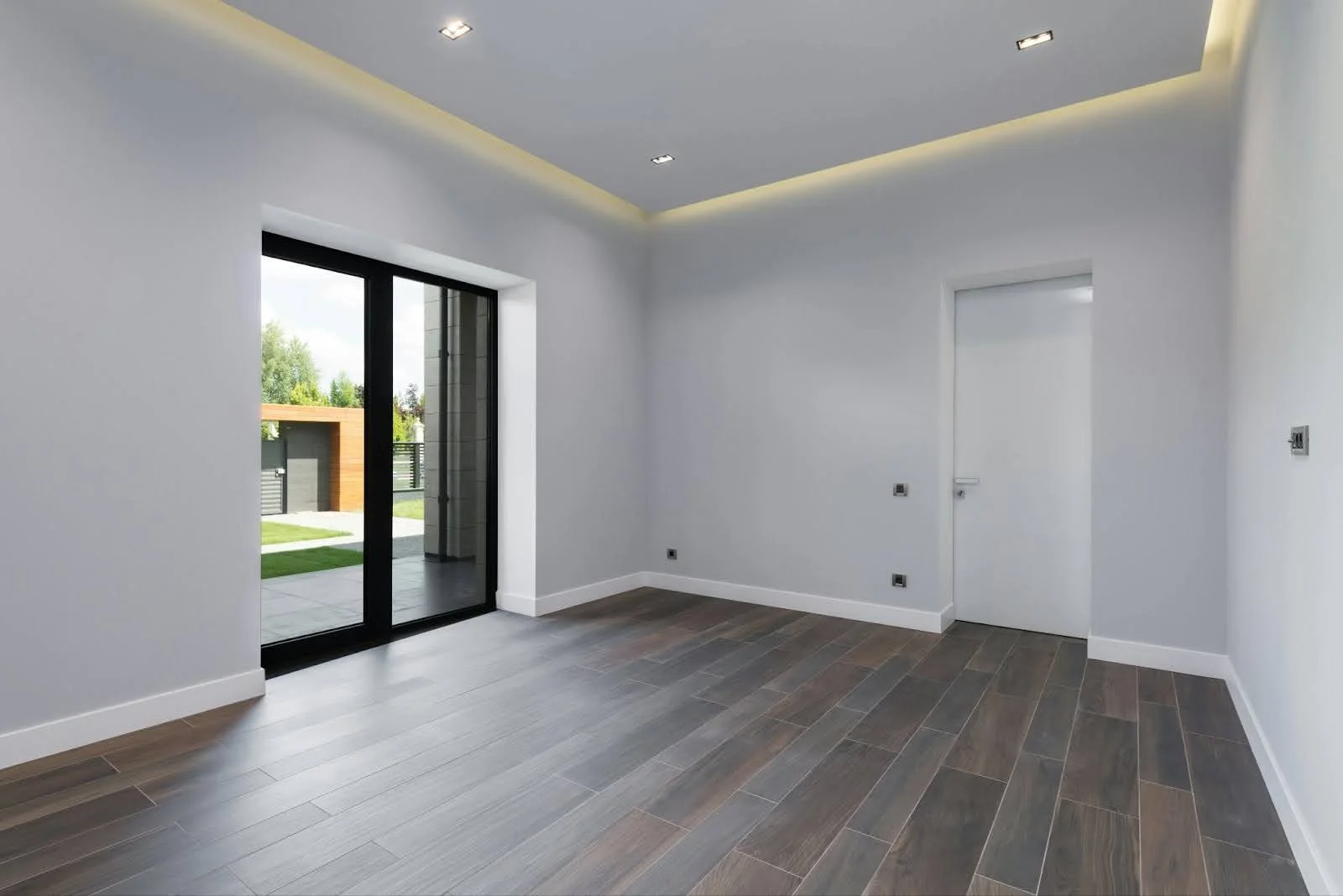 picture of a minimalist empty room featuring dark wood look floor tiles a white door and large black framed glass sliding doors leading to a courtyard