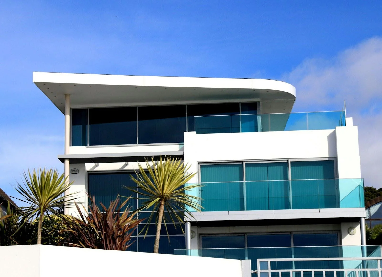 picture of a modern white multi level home featuring floor to ceiling glass windows and balconies with glass railings against a clear blue sky