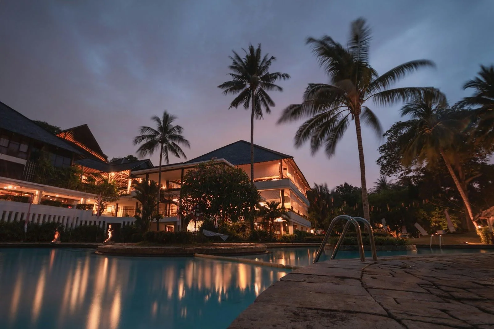 picture of a resort style pool at dusk reflecting a large multi story building with warm interior lighting and palm trees
