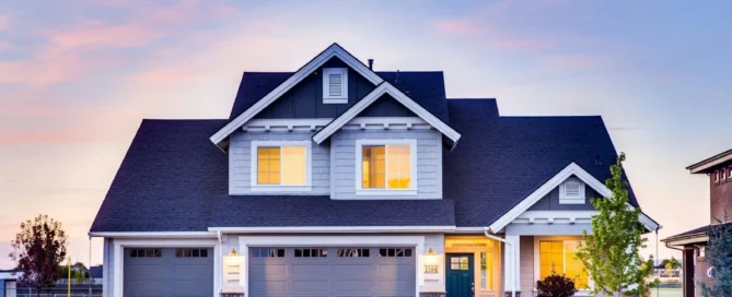 picture of a two story luxury home remodel blue house with white trim and three garage doors viewed at dusk with warm lights glowing in the windows