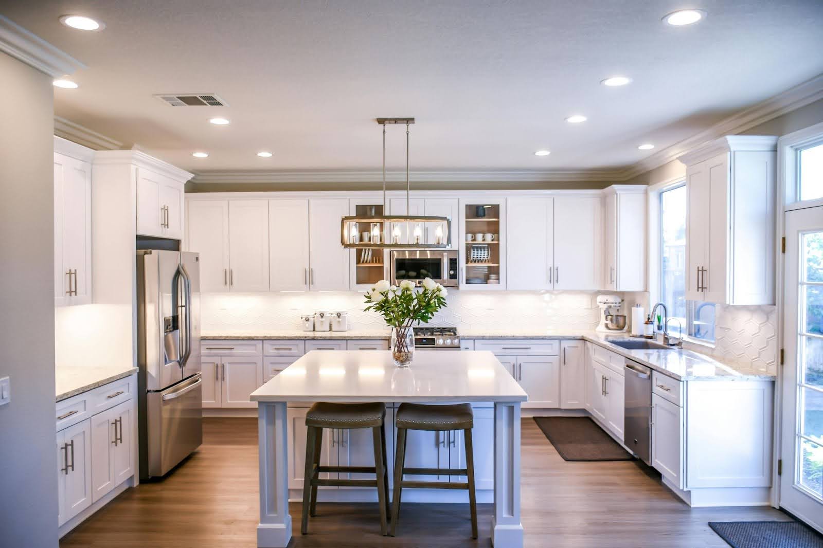 picture of an expansive white kitchen featuring a large square island with a flower vase stainless steel appliances and a rectangular glass chandelier
